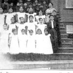 Staff and children in uniform posed on the front steps of Ahousaht. Image and caption is cropped, caption reads: 