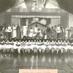 Children in a Christmas play on stage with a choir seated below them in large gymnasium, caption reads Christmas 1963 - Portage la Prairie