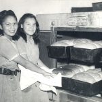 Two people standing to one side, pulling a rack of bread loaves out of oven at Portage la Prairie Residential School.