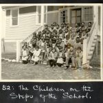 Large group of children sitting on the steps of the school. Caption below reads: 