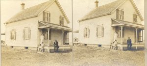 One person standing and two people sitting on the porch of a house, photographed to capture image of full building.