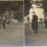 One person standing next to a camera on a tripod. Two other people standing together away from photographer. Outdoors, grass, trees and building seen in distance.