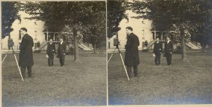 One person standing next to a camera on a tripod. Two other people standing together away from photographer. Outdoors, grass, trees and building seen in distance.