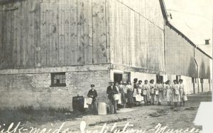 Children lined up, holding milk buckets outside along the wall of a barn.