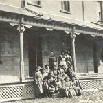 Children and staff, seated and standing posed for a photo on the steps of the teacher's residence and hospital.