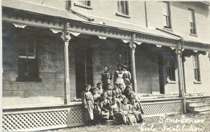 Children and staff, seated and standing posed for a photo on the steps of the teacher's residence and hospital.