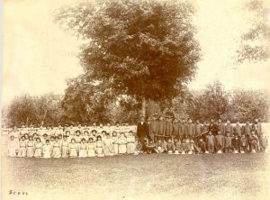 Group of youth, divided by assumed gender seated and standing for a photograph outdoors under a tree.