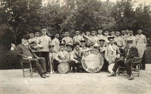 Children in uniforms holding instruments, standing and crouched with two staff members in front seated in chairs, all posed for a photograph outdoors.