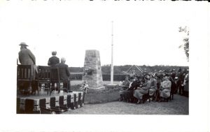 Crowd sitting in chairs, outdoors with large stone cairn in front of them, some individuals seated on a stage..
