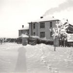 File Hills Residential School building in winter, snow on the ground, smoke coming out of the chimney, taken from across the lawn, large sign at the front of the building.