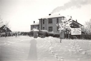 File Hills Residential School building in winter, snow on the ground, smoke coming out of the chimney, taken from across the lawn, large sign at the front of the building.