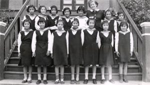 Group of youth in Canadian Girls in Training uniforms, standing posed for a photograph outdoors on the stairs of File Hills Residential School, with Mrs. Lachlan Mclean, the principal's wife.