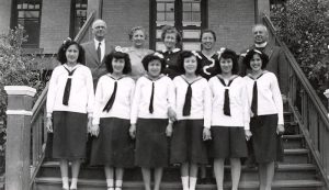 Six youth in Canadian Girls in Training Uniform standing posed for a photograph outside on the stairs of File Hills Residential School, with a group of staff posed behind them.