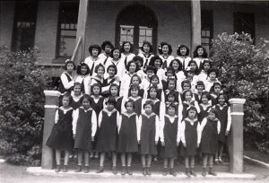 Large group of youth in Canadian Girls in Training Uniforms, standing posed for a photograph on the front steps of File Hills Residential School.
