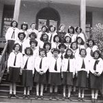Group of youth in Canadian Girls in Training uniforms standing posed on the front steps of File Hills Residential School, with Mrs. Lachlan McLean in the back row.