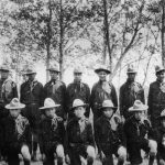 Group of youth in boy scout uniforms kneeling and standing posed for a photograph outdoors in front of a line of trees.