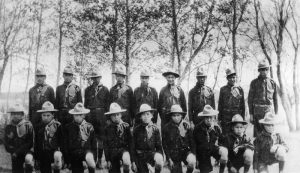 Group of youth in boy scout uniforms kneeling and standing posed for a photograph outdoors in front of a line of trees.