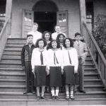 Group of youth with two staff members standing posed in uniform on the front steps of File Hills Residential School.