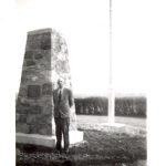 Individual standing in front of large stone cairn with flagpole and line of trees in the background.