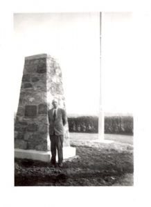 Individual standing in front of large stone cairn with flagpole and line of trees in the background.