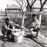 Two children squatting, posed for a photograph with paint buckets in front of a fence, some buildings in the background.