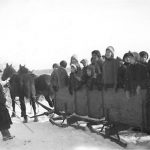Group of youth standing in a large box sleigh, two horses pulling it and two individuals beside the sleigh.
