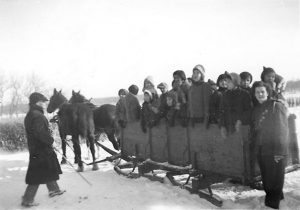 Group of youth standing in a large box sleigh, two horses pulling it and two individuals beside the sleigh.