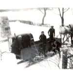 Two staff members on a box sleigh with two children ready to drive, and a team of two horses. Large stone cairn in the background.