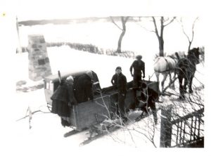 Two staff members on a box sleigh with two children ready to drive, and a team of two horses. Large stone cairn in the background.