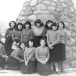 Group of youth sitting and standing in front of a large stone cairn, outdoors in winter.