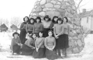 Group of youth sitting and standing in front of a large stone cairn, outdoors in winter.