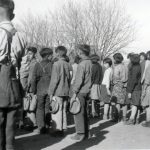 Children standing in lines, some with backs to the camera, some looking at the camera, watching flag being raised (flag not pictured).
