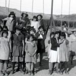 Group of children, some wearing hats, standing posed for a photograph on and in front of a swing set.