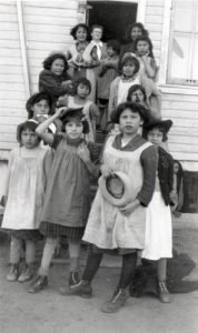 Students wearing and holding hats, standing with a staff member on steps with Residential School in the background.