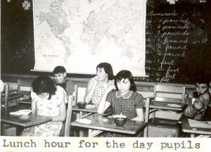 Children at desk eating with following text underneath: Lunch hour for the day pupils.