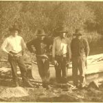 Four Cree people stand on the shoreline, A canoe tied on the water directly behind them. Paddles are seen resting in the canoe and on the ground by the people's feet.