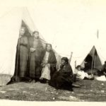 Cree people seen standing at the entrances of their shelters.