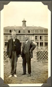 The two reverends wearing suits standing on the lawn for a portrait with Norway House Residential School seen in the background.