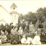 Visiting reverend standing with members of the congregation in front of the church building for a posed group photograph.