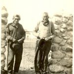 Two elderly people standing outside at the entrance of a stone fence seen directly behind them.