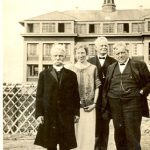 Visiting reverends standing with an unidentified person. Norway House Residential School Building is seen in the background.