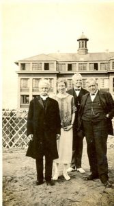 Visiting reverends standing with an unidentified person. Norway House Residential School Building is seen in the background.