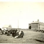 Candid photograph of the parents at their camp. The Norway House Residential School Building is seen in the background of the camp.