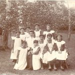 Ten children in aprons posed around a staff member in yard