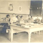 Sewing room with sewing machines and a large table with sewing materials, children and staff sitting behind large table