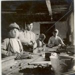 Group of four children standing at a workbench using carpentry tools, indoors, some looking at the camera.