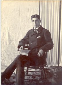 Formal portrait of Tommy Keefer, seated holdin ga book, wearing a suit, backdrop and curtains in the background.