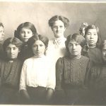 Formal portrait of youth, wearing uniforms with staff of the school, taken inside.