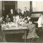 Children in uniform seated for dinner, with staff members standing behind, all looking at the camera.