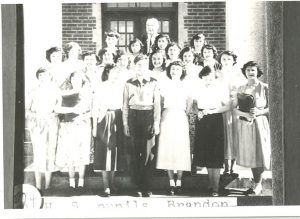 Group of high school youth with Rev. Oliver B. Strapp, standing posed for a photograph in front of Brandon Residential School.